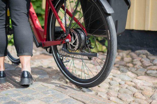 A closeup at a black kickstand on a red bike, you can see that a woman is walking with the bicycle