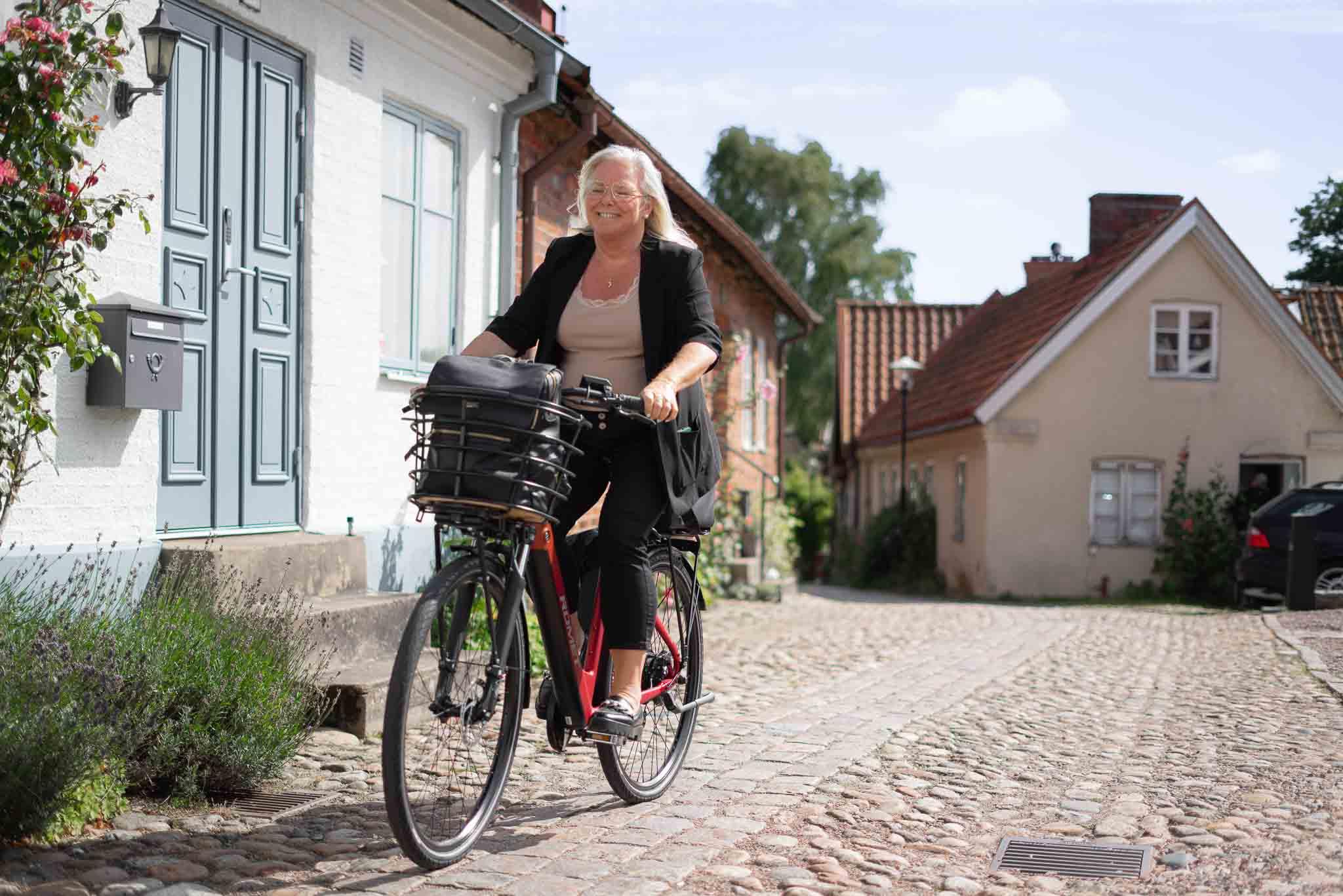 A woman bikingin an old town, she has a bag in her curved basket and she looks happy