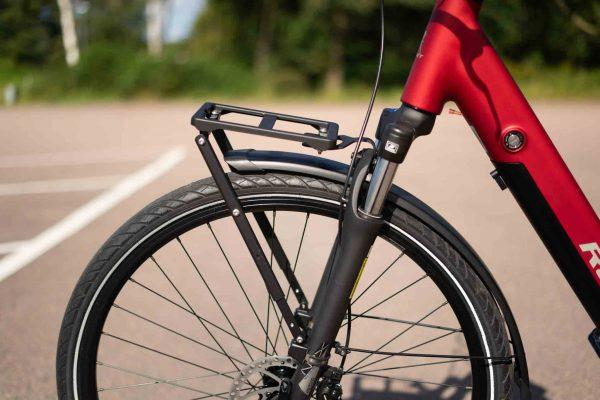 A front rack on a red bicycle, with asphalt and a forest visible in the background.