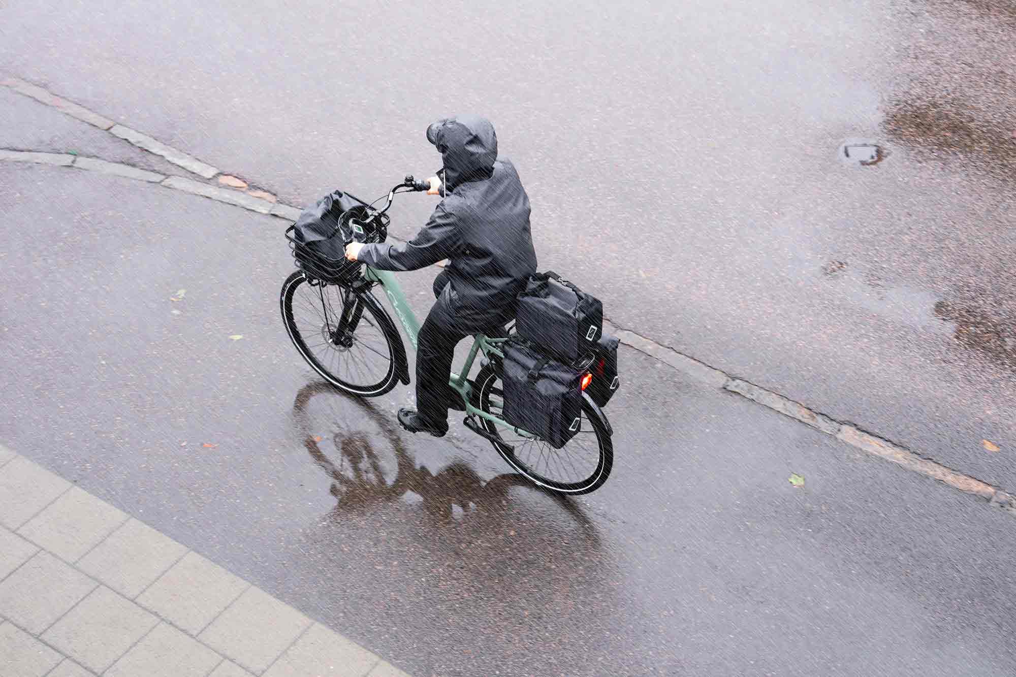 A woman biking in the rain with a front top bag, a top bag in the back and two side panniers. They are all wateproof and grey.