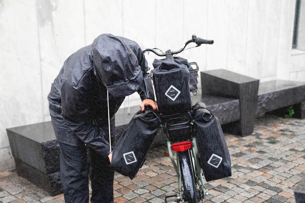 A woman lifitng up a grey sidepannier from her bicycle in the rain.