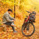 A man sitting on a bench in a yellow autumn forest with his bicycle infront of him