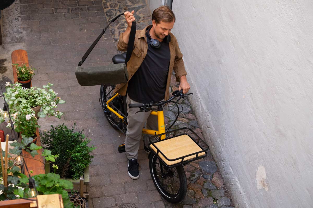 A picture of a man with a green bag and a yellow bicycle in an alley