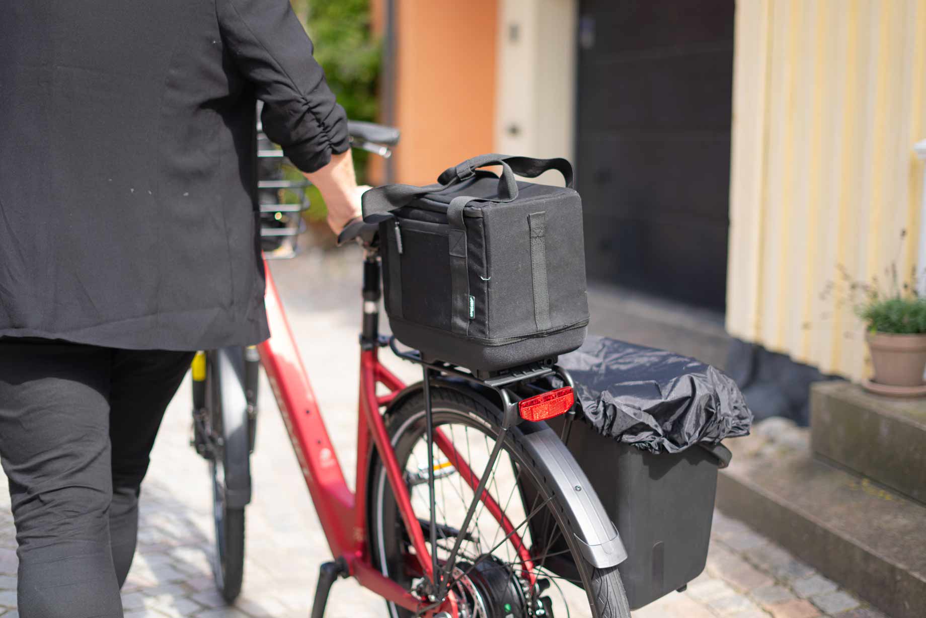 A red bicycle with a lady walking on a street. On the bicycle there is oine top bag and a sidecrate