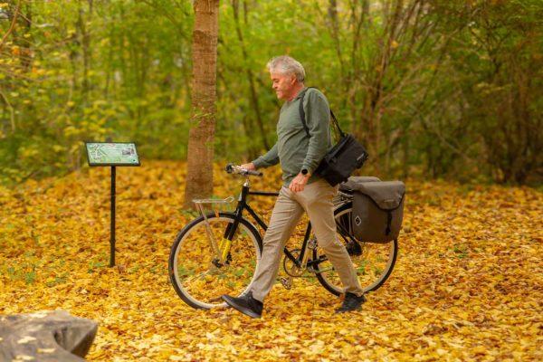 A man walking in a yellow autumn forest with his bicycle. On his shoulder he has a small bicycle bag.