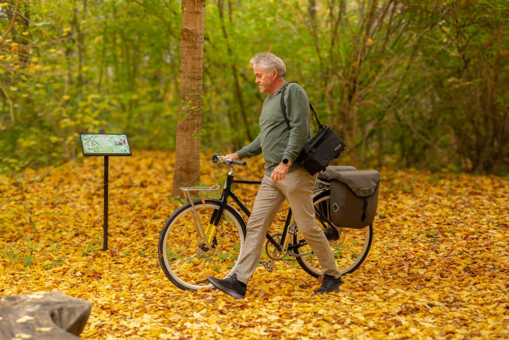 A man walking in a yellow autumn forest with his bicycle. On his shoulder he has a small bicycle bag.