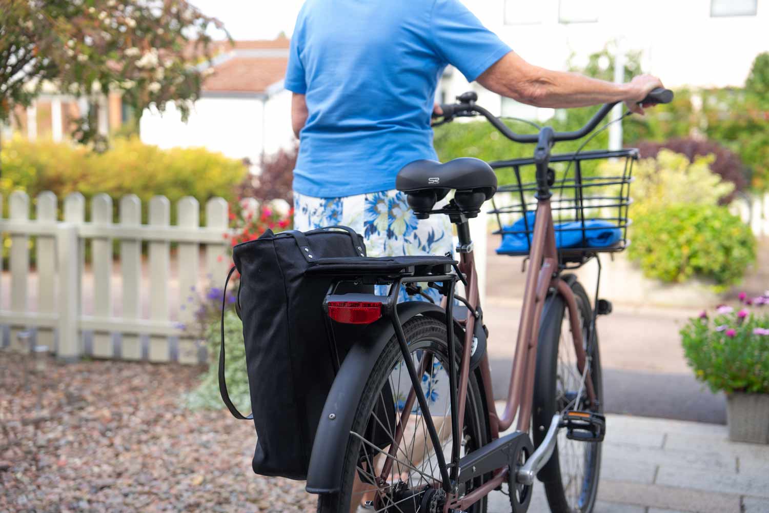 A lady walking with her bike in her front yard with a black bag on the side of the rear carrier