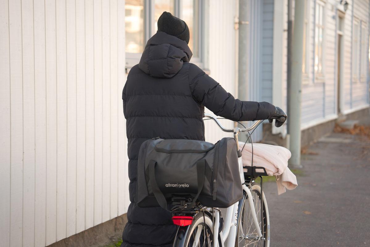 A woman walking in the winter with a bicycle. On the rear carrier she has a big sporty bag.