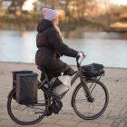 A girl biking by a river with two bags on the rear carrier