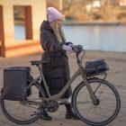 A girl walking with her bike by a river with two bags on the rear carrier