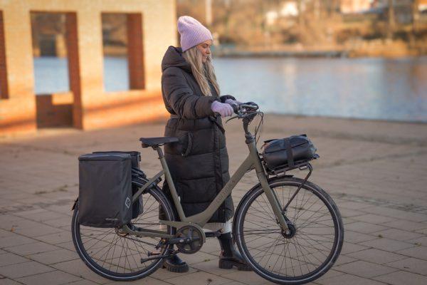 A girl walking with her bike by a river with two bags on the rear carrier