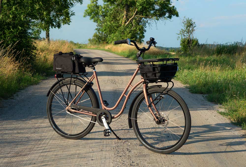 A bicycle in the middle of a gravel road. On the front there is a basket and in the back there is a small top bag.