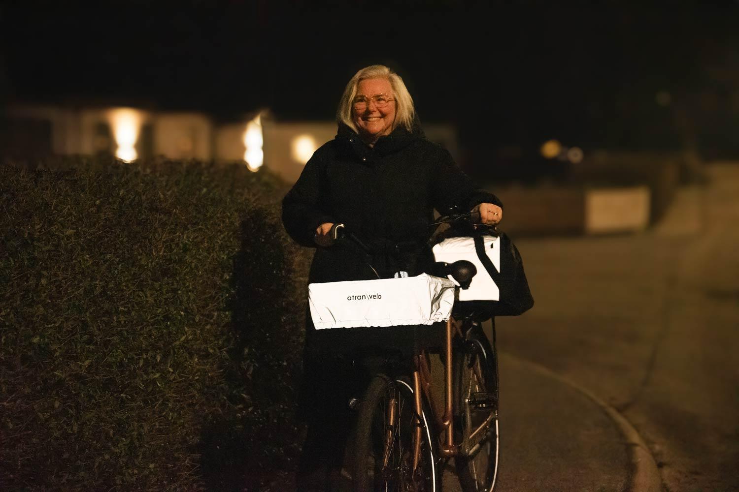 A woman walking with her bike in the dark. On the front she has a reflective cover on her basket and in the back she has an reflective bag.