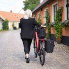 You see a woman dressed in black walking with a bicycle in an old, beautiful city, the bike have a bag and a crate.