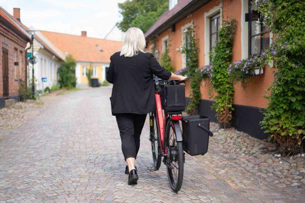 You see a woman dressed in black walking with a bicycle in an old, beautiful city, the bike have a bag and a crate.