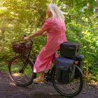 A woman biking with three bags on her rear carrier and a basket in the front, outside in a forrest.