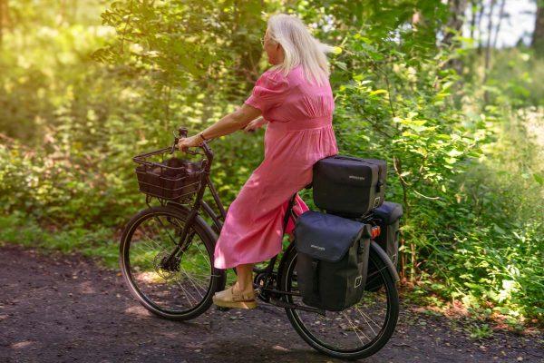 A woman biking with three bags on her rear carrier and a basket in the front, outside in a forrest.