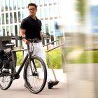 A man walking with his grey bicycle. On the rear carrier he has two black sidepanniers.