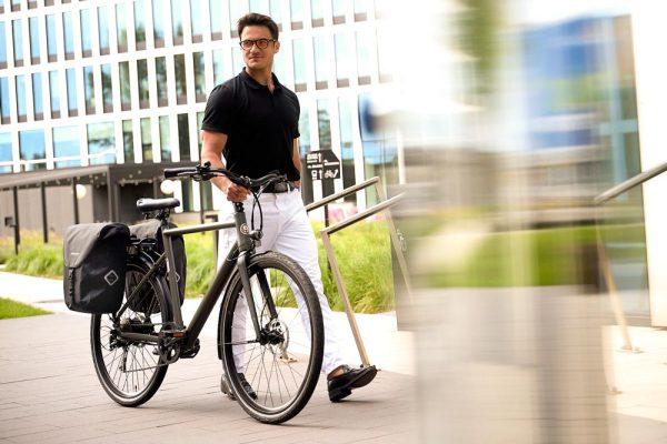 A man walking with his grey bicycle. On the rear carrier he has two black sidepanniers.