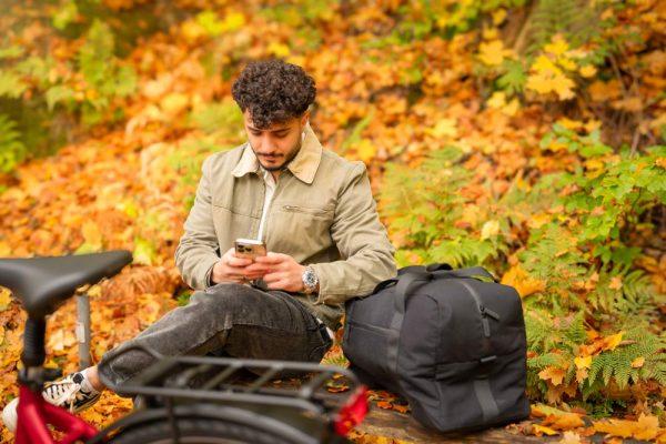 A man sitting on a bench in a yellow forest, he has his elbow on a big bicyclebag. In the front of the picture you see a blurred carrier.