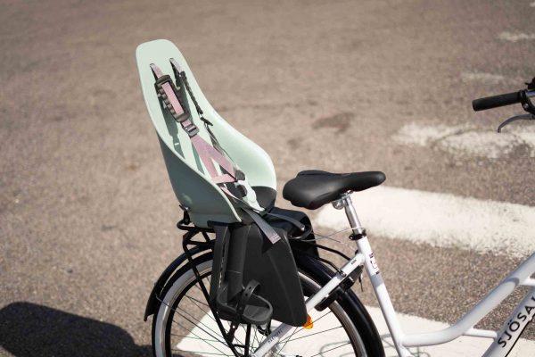 A white bicycle with a black rack at a crosswalk with a childseat on top.