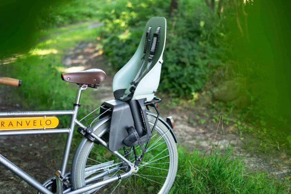 A black rack on a trail with greenery all around. And a childseat on the carrier.