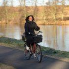 A happy girl biking by a river and on her bike she has a black basket in the fron and a netbasket in the back