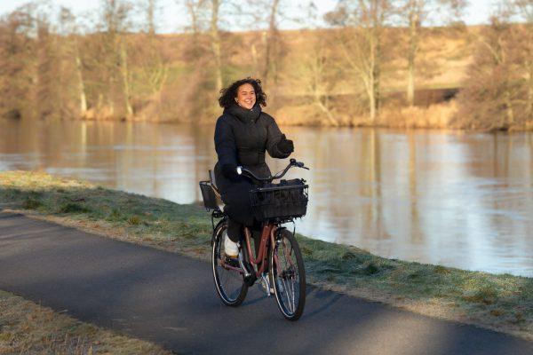 A happy girl biking by a river and on her bike she has a black basket in the fron and a netbasket in the back