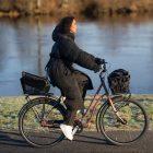 A girl biking alongside a river, she has two different baskets, one at the front and one in the back