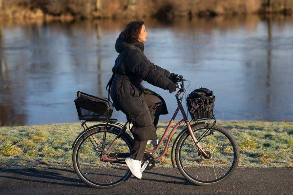 A girl biking alongside a river, she has two different baskets, one at the front and one in the back