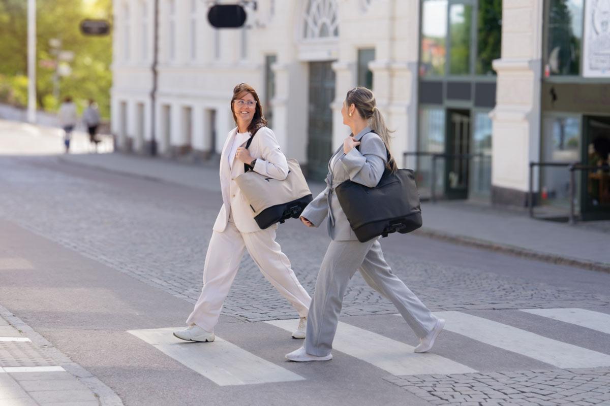 Two business women walking across a crossing with one stylish and feminine bag each. One in black and one in beige.