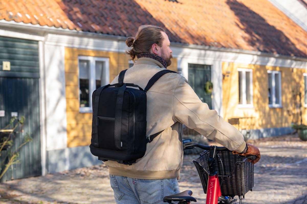 A lifestylepicture of a man walking with his bike from behind with the black metro backpack on his back, the area is an old town